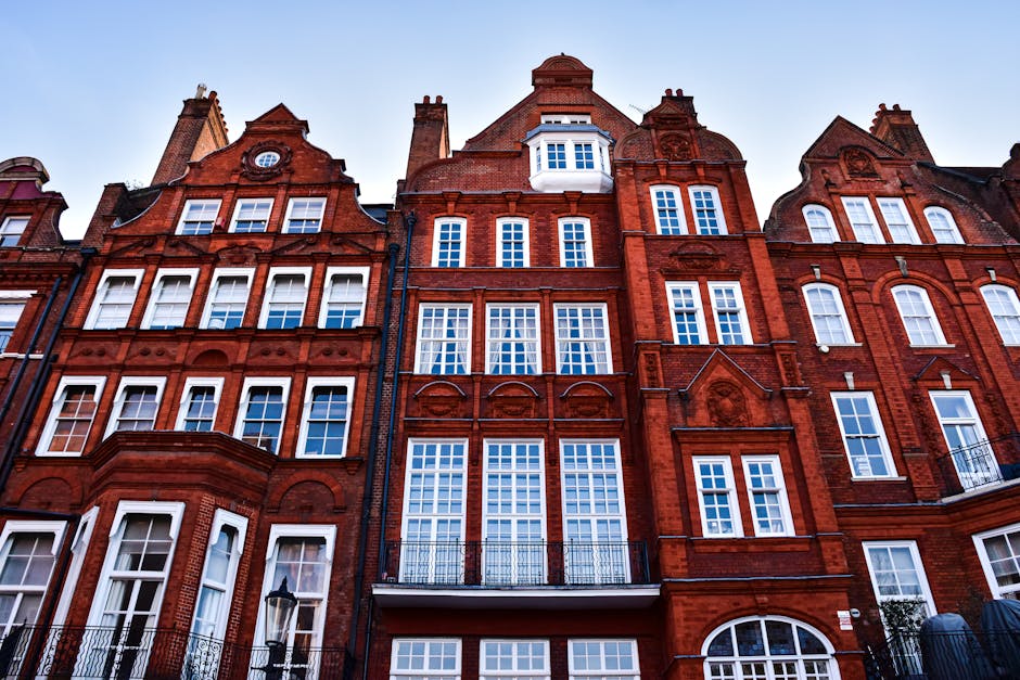 A row of historic red-brick townhouses in Knightsbridge, SW1X, featuring multiple windows with white frames, some with decorative arches and small balconies. The buildings have ornate architectural details, including stone carvings and gabled roofs, with the lower floors showing a doorway and ground-level windows. The street-facing facades are well-lit by natural daylight, capturing the height and grandeur typical of London’s residential architecture. In front of the buildings, a section of pavement is visible, along with a traditional street lamp. This image exemplifies the type of property that [COMPANY_NAME], Man With a Van Knightsbridge, specializes in for home relocation, furniture transport, and packing and moving services, involving careful handling of furniture, boxes, and other household items during the loading process into a moving van for interior or exterior transportation.